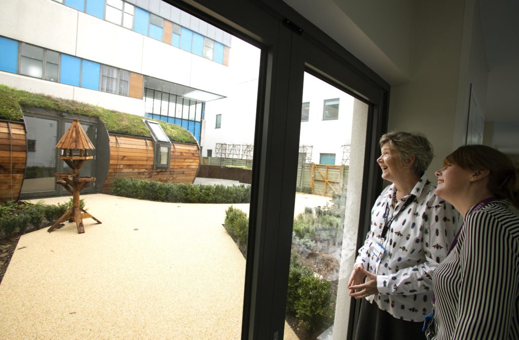 Clare Curran and Barbara Buckley looking out at the Time Garden through the glass door from the corridor
