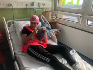 A picture of Fred, wearing a hi-vis jacket and pink builders helmet, sat on his hospital bed overlooking the building site where the breast care centre is being built