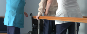 A person doing rehab practicing walking  while holding on to a railing. There is a nurse beside and next to them offering support.