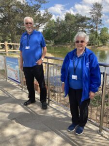 Peter and Helen Mockford wearing blue volunteer tshirts standing by a pond