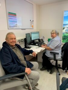 man sitting in clinic room with woman in clinical tunic at computer. Both are looking at camera