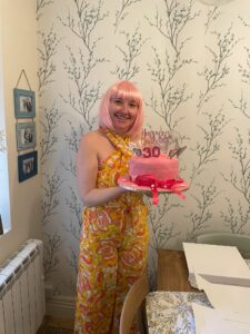 Woman with pink wig wearing yellow jumpsuit and holding pink birthday cake with 30 on it