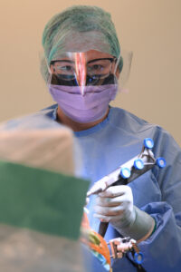 doctor in full theatre gown, mask and visor looking intensely with a tool in her hand