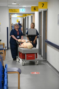 two nurses pushing a machine with a mannikin along a hospital corridor