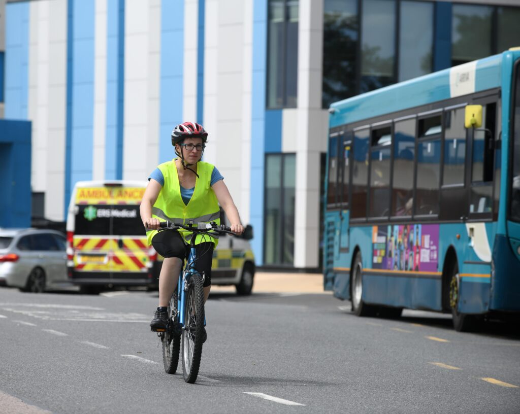 A woman is riding a blue bicycle along a road with an ambulance, car, bus and blue and white stripy building in the background. She is wearing a t-shirt, short trousers, a cycling helmet and a high visibility yellow vest.