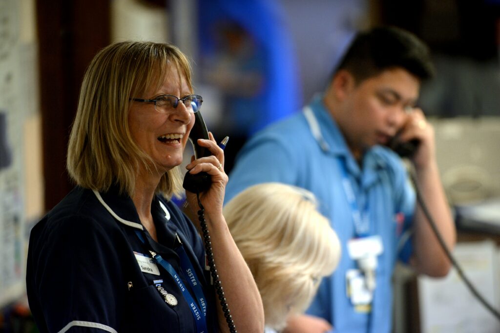 A woman and a man stand next to each other both holding telephone handsets. They are wearing NHS uniforms. The woman is in the foreground and wearing a dark blue uniform including a fob watch. She is smiling. The man is in the background and wearing a sky blue uniform. Both the man and the background are blurred, as the focus is on the woman.