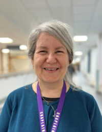 Sarah Amos standing in a hallway with a purple lanyard around their neck, smiling at the camera.