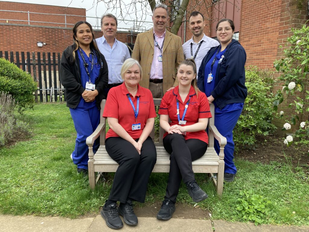 A group of people in various clinical uniforms pose for a photograph. Two people are sat on a bench with the remaining five people stood around them.