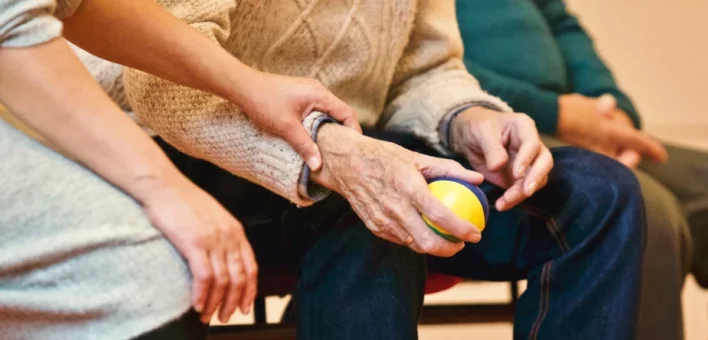A carer's hand holds a patient's hand. The patient has a ball in their right hand.