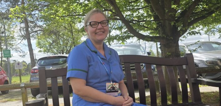 Woman in nursing uniform sitting on bench under a tree. Smiling to camera.