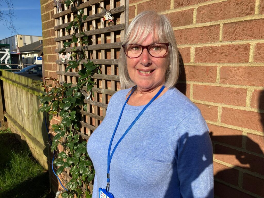 A woman with grey hair and glasses, wearing a sky blue jumper, stands smiling with a wall in the background.