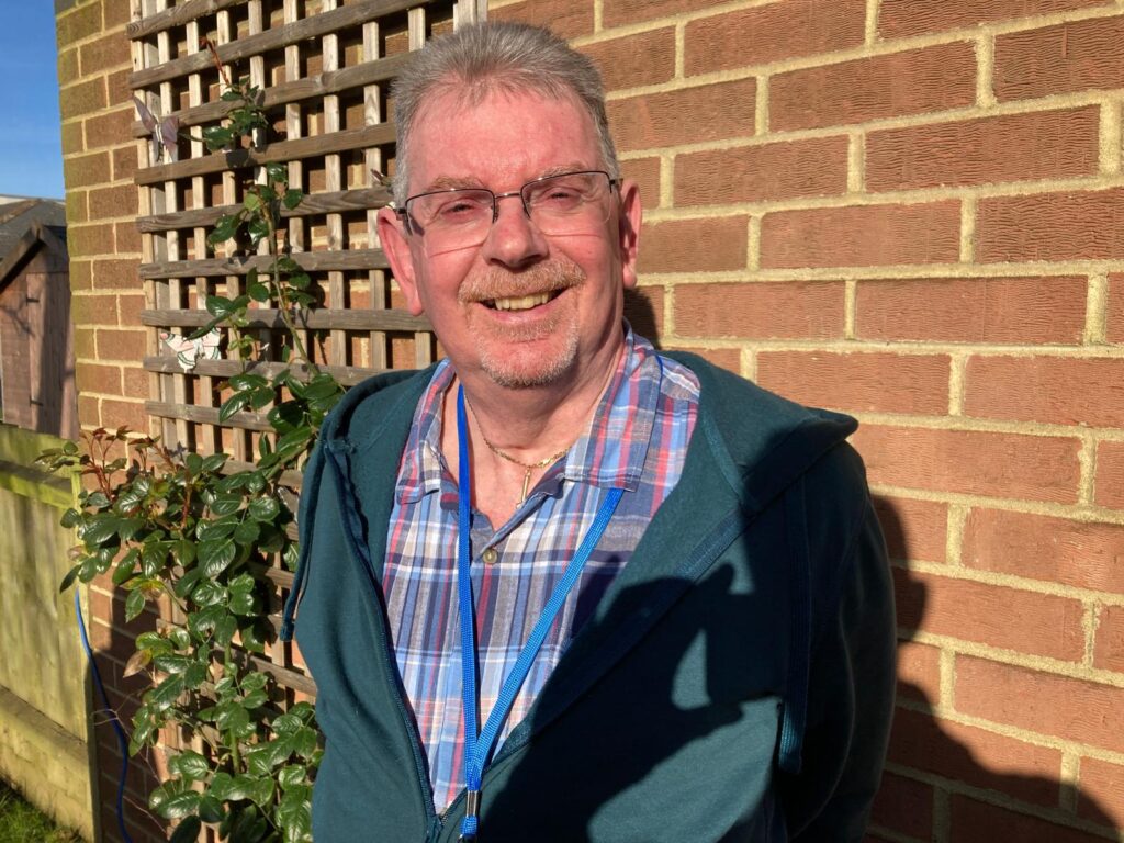 A man with grey hair and a grey moustache stands in front of a wall, wearing glasses, a green hoodie and checked shirt. He is smiling at the camera.