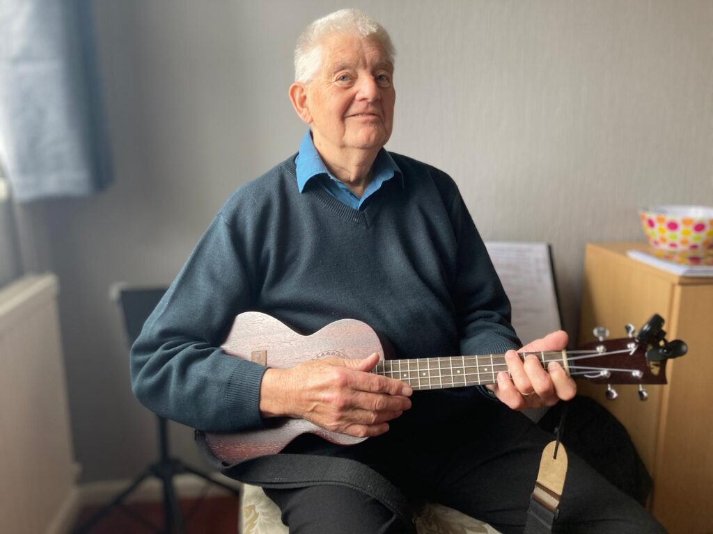 A man sits in a chair, smiling at the camera, playing a ukelele