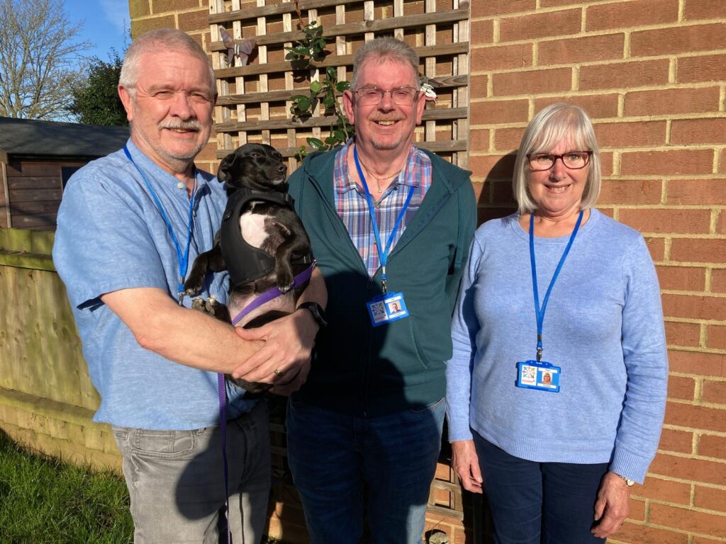 Three people stand in a row, against a wall. The man on the left, Terry, is holding a small black dog.They are all smiling at the camera.