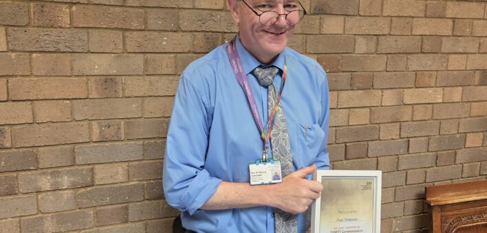 Paul holds his framed certificate and smiles at the camera